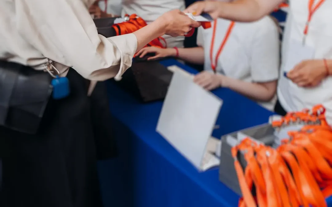 Two people exchange an event badge at a registration desk with a laptop and orange lanyards.